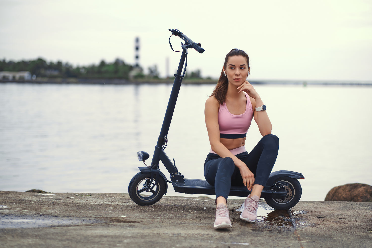 En kvinna i träningskläder sitter på sin parkerade elsparkcykel med havet i bakgrunden